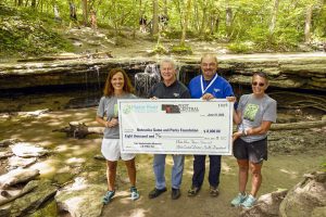 Four people hold an oversize check standing in front of a waterfall in a wooden area in Eastern Nebraska.