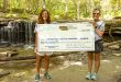 Two women hold a large fake check while standing in front of a waterfall in a wooded area in eastern Nebraska.
