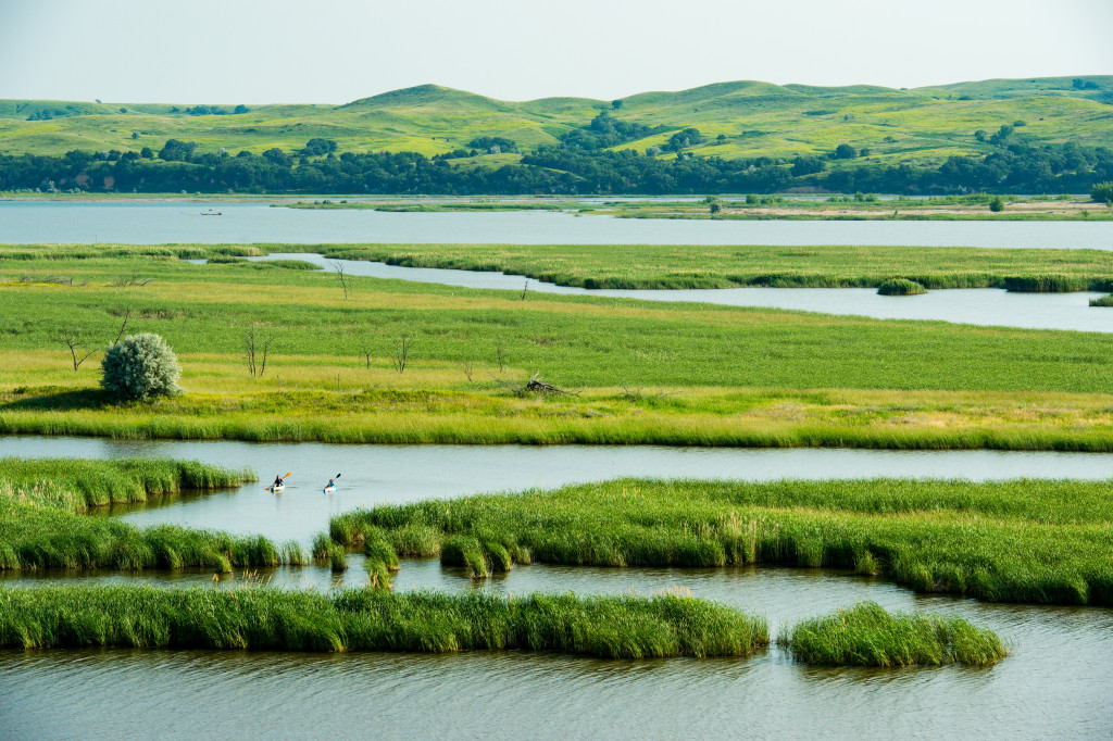 Two women kayak in a meandering backwater of a river with grass-covered sand dunes, known as the Sandhills, in the background.
