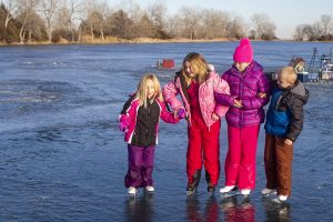 Three young girls and one boy skate gingerly on a frozen lake.