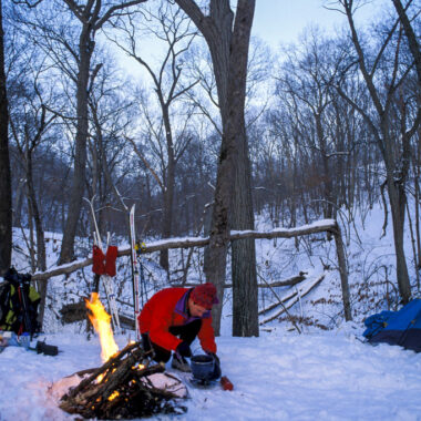 A man at a snowy campground with his tent, gear and a campfire during winter.