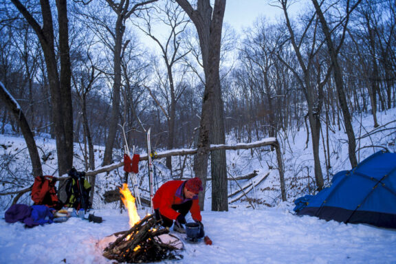 A man at a snowy campground with his tent, gear and a campfire during winter.