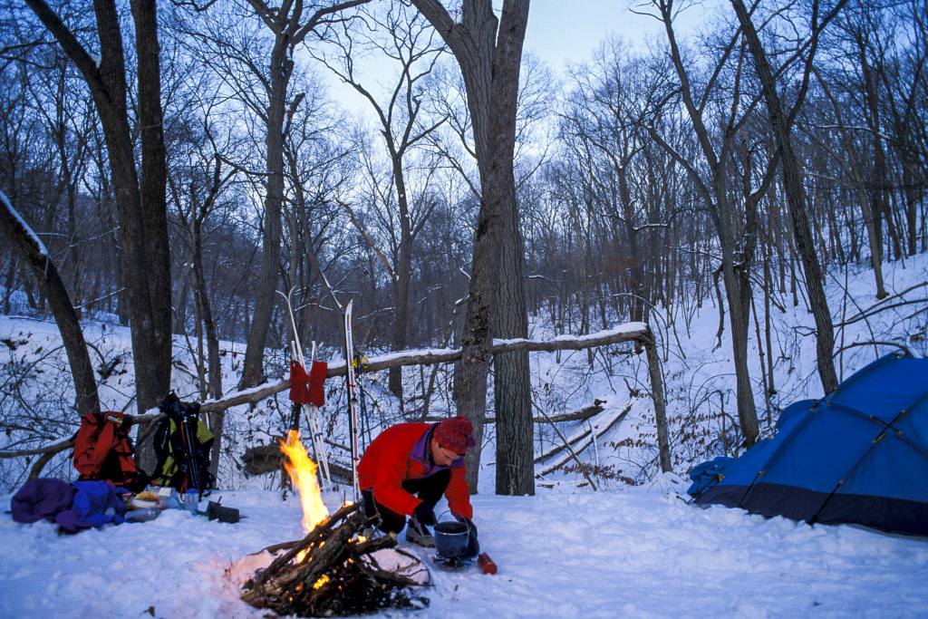 A man at a snowy campground with his tent, gear and a campfire during winter.