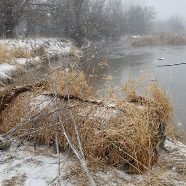 A bale of hay along a snowy river bank during winter as snow falls.