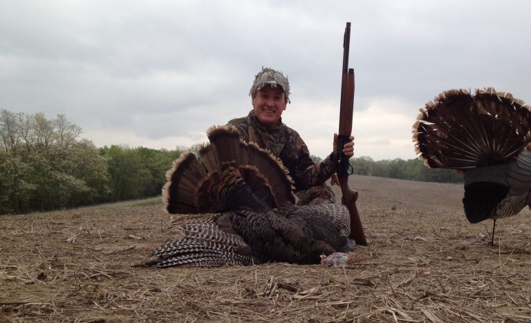 A man poses in a field with a harvested turkey and hunting decoy.