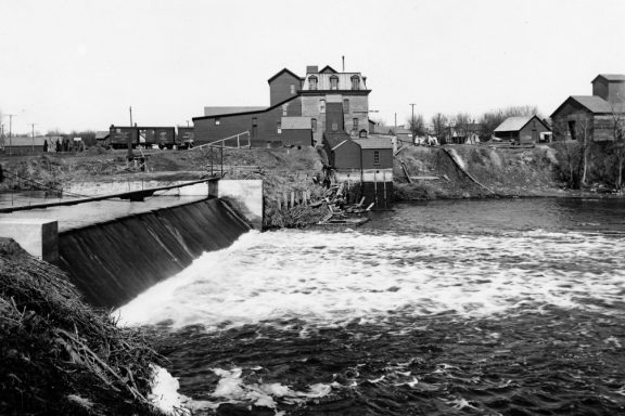 Water pours over a dam, a flour mill in the background