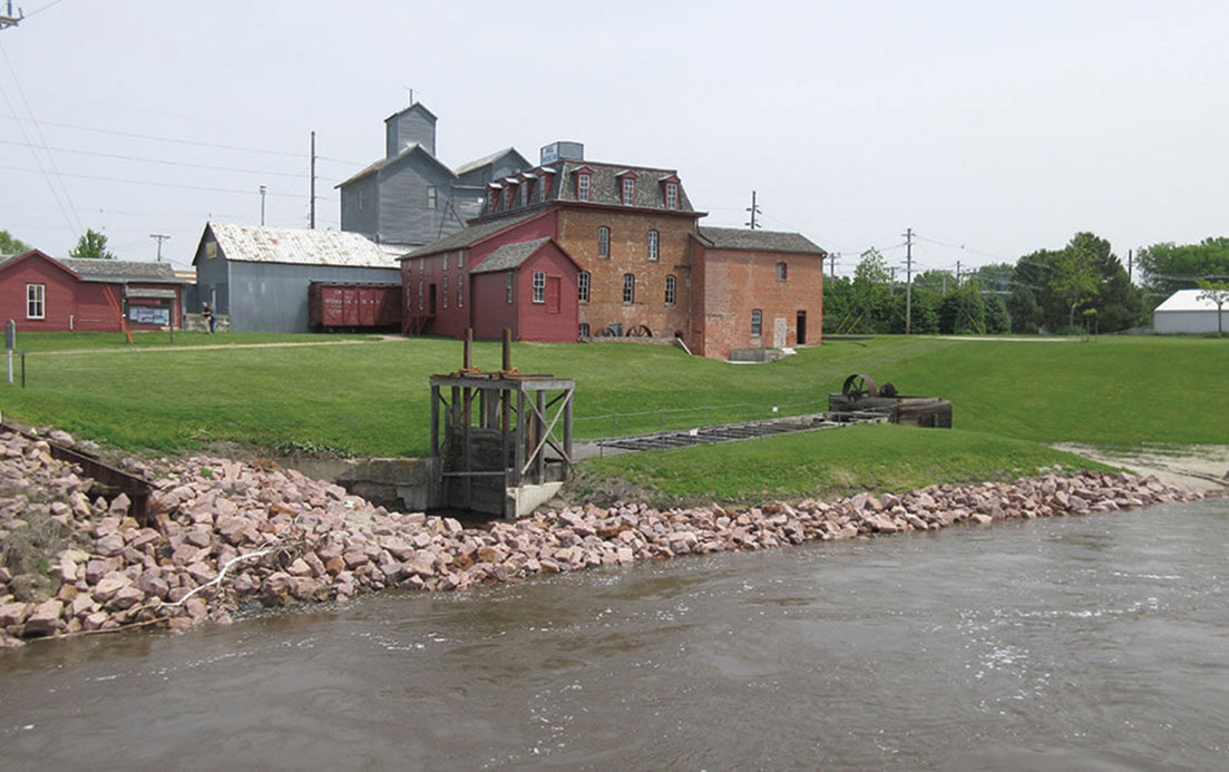 A color photograph of a shallow river, a flume and mill in the background