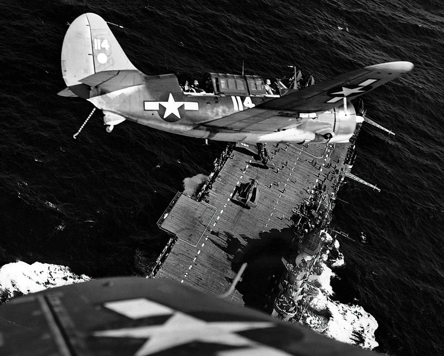 A black and white photo captures a Helldiver airplane turn  its nose down to an aircraft carrier below floating on a calm ocean