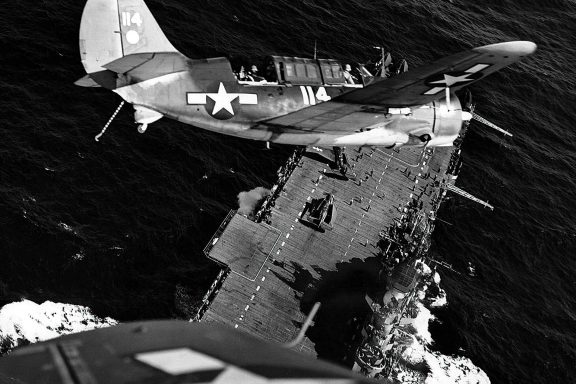 A black and white photo captures a Helldiver airplane turn its nose down to an aircraft carrier below floating on a calm ocean