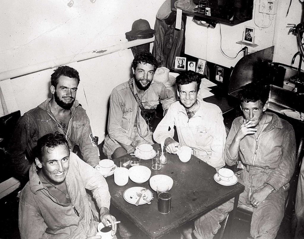 Four soldiers, unwashed, sit and smoke around a small square dinner table in a military tent