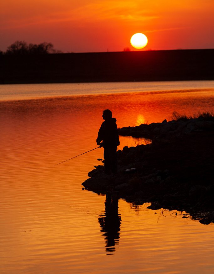 A man fishes during a red summer sunset on the back of a lake.