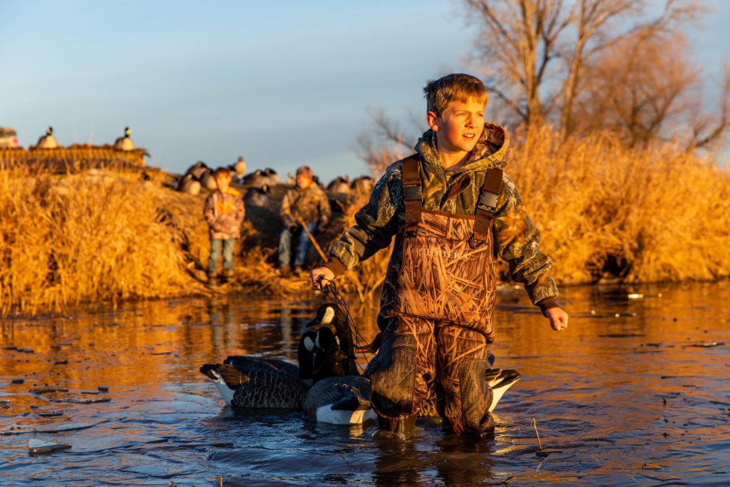 A boy wads in shallow water and holds goose decoys while goose hunting.