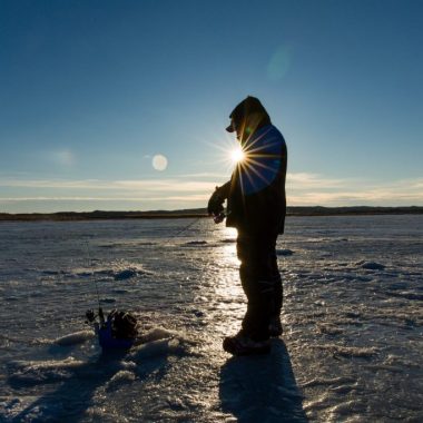 A man stands on a frozen lake preparing to ice fish with the sun beaming behind him.