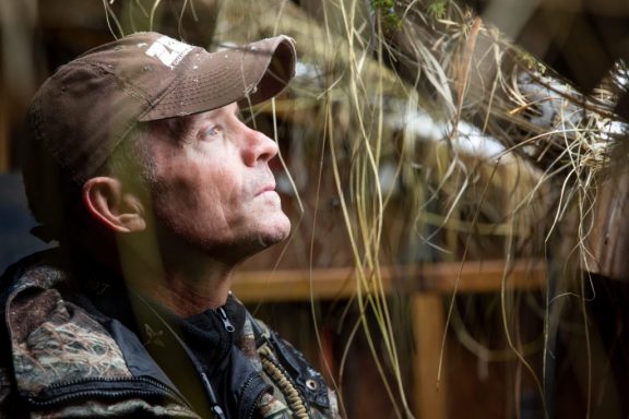 A man inside a waterfowl hunting blind looking out.