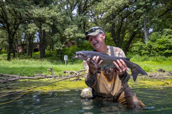 A man wearing waders in a lake smiles while holding a catfish he caught.