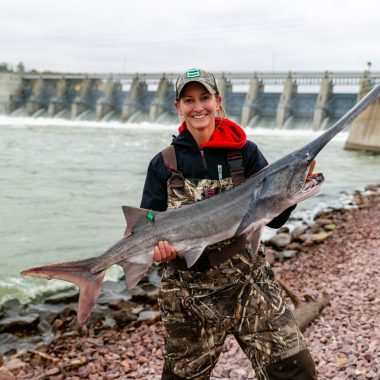 A woman smiles and holds a large paddlefish she caught near Gavins Point Dam.