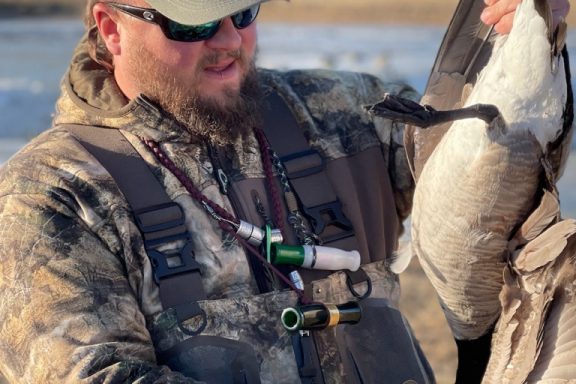 A man wearing duck calls around his neck holds a goose he harvested