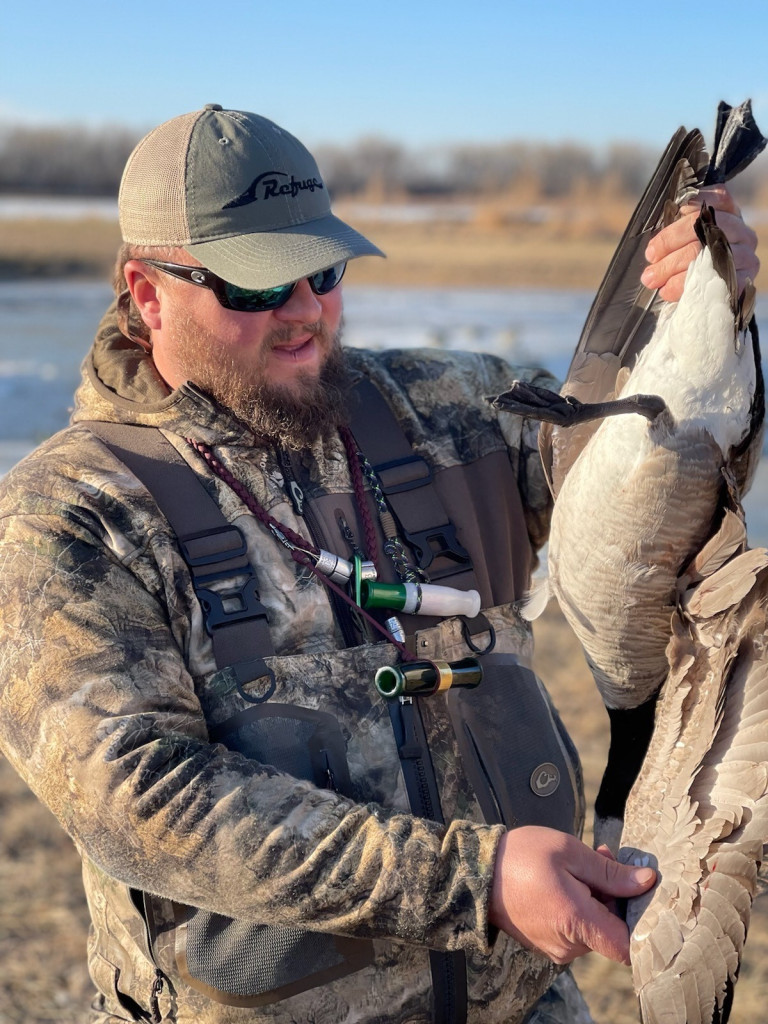 A man wearing duck calls around his neck holds a goose he harvested