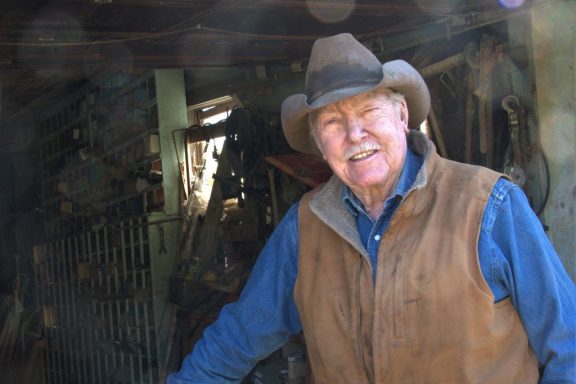 Wearing a blue, long-sleeved shirt, brown vest and beige cowboy hat, Paul Allen stands in the doorway of his barn-looking shop.