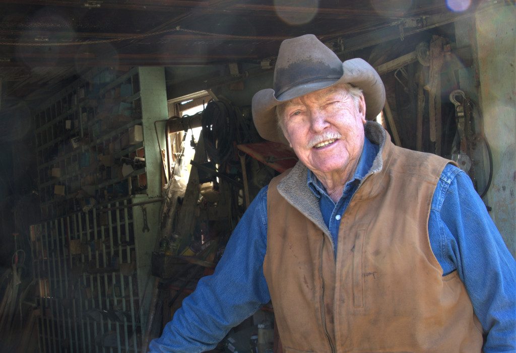 Wearing a blue, long-sleeved shirt, brown vest and beige cowboy hat, Paul Allen stands in the doorway of his barn-looking shop.