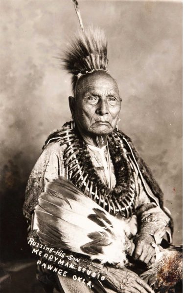 A black and white portrait of a Native American man in traditional dress, holding ritual items