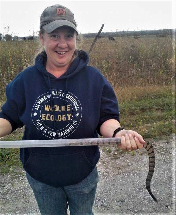 A woman holds a tube with a snake in it; she hangs onto its tail