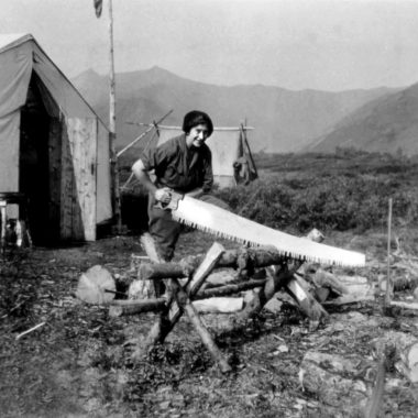 A woman in men's work clothes uses a large handsaw to cut wood