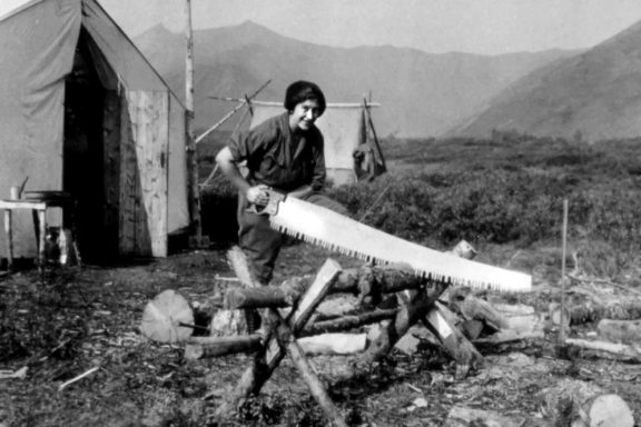 A woman in men's work clothes uses a large handsaw to cut wood