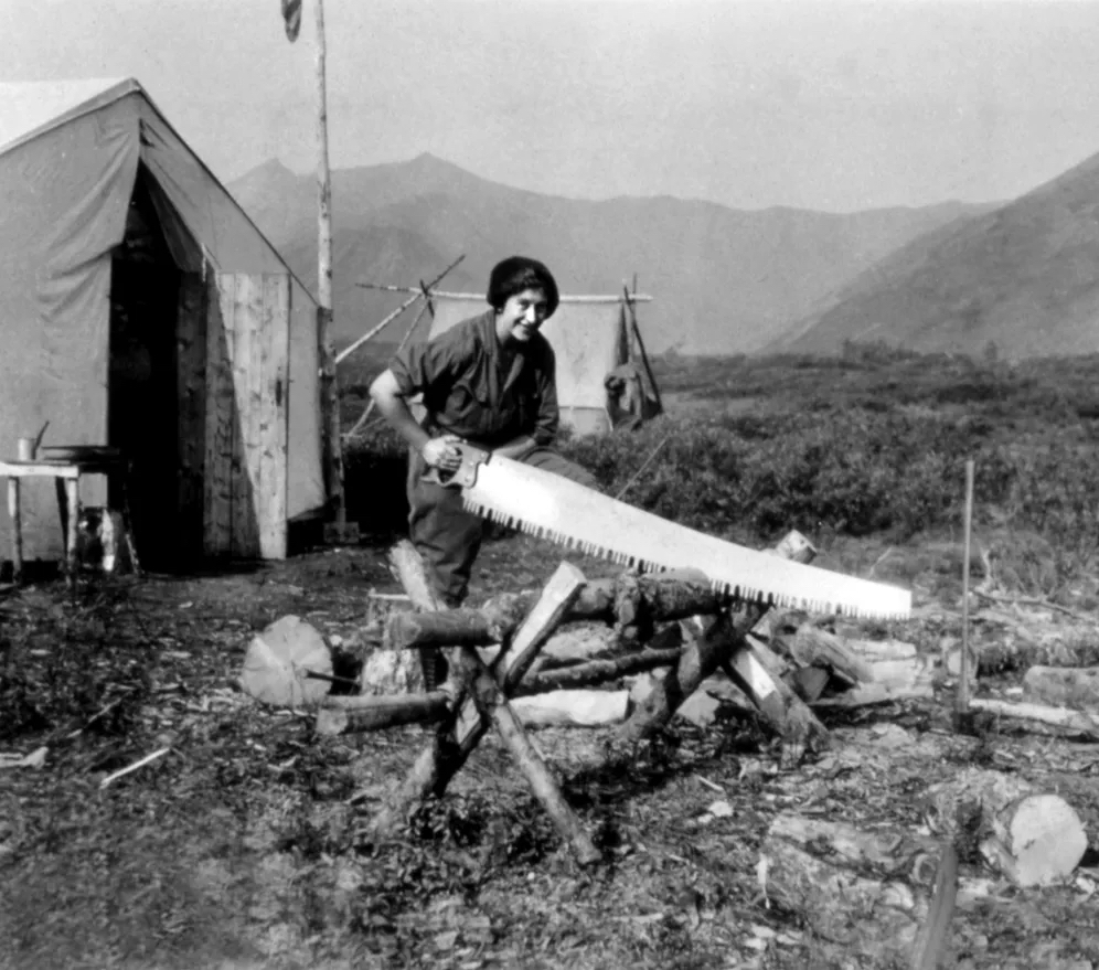 A woman in men's work clothes uses a large handsaw to cut wood