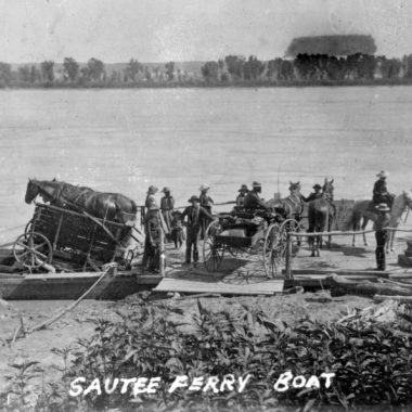 a rickety plank ferry with paddlewheels is loaded with wagons and carts