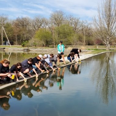 a row of students crouches by a waterbody to put their trout fish in