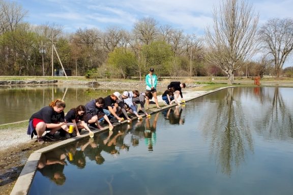 a row of students crouches by a waterbody to put their trout fish in