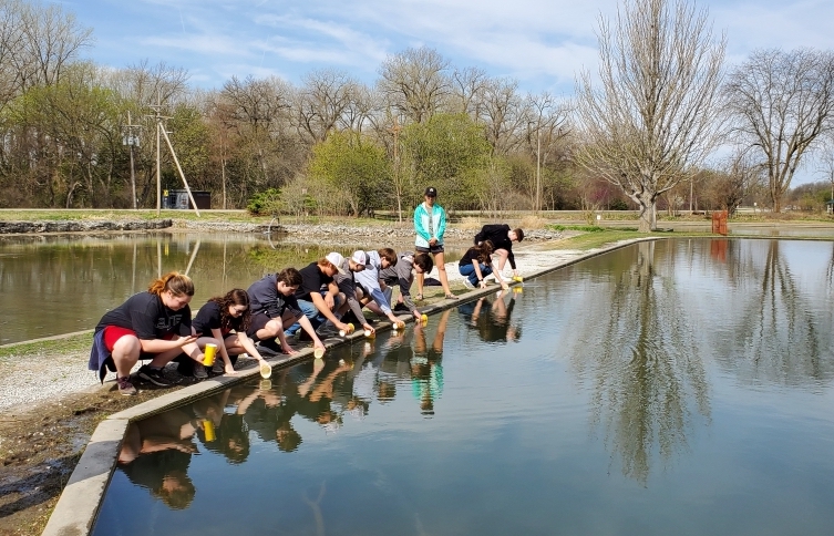 a row of students crouches by a waterbody to put their trout fish in