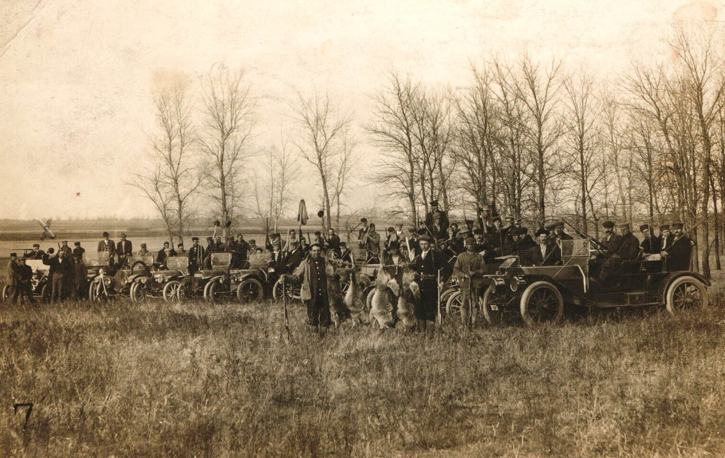 a row of men on foot and in vehicles stands at a tree edgeline with their firearms