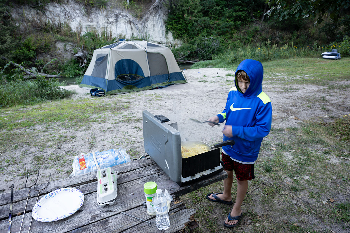 a boy cooks eggs in a skillet at a tent campground