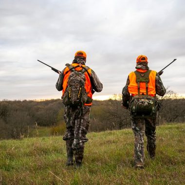 Two deer hunters walk along a tree line.
