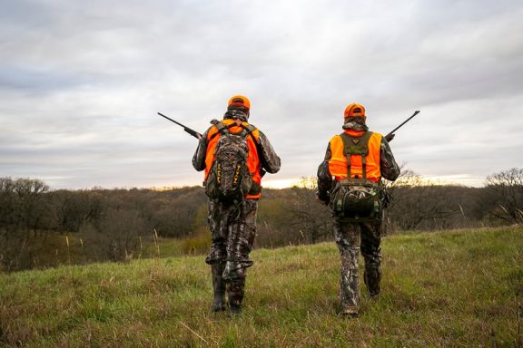 Two deer hunters walk along a tree line.
