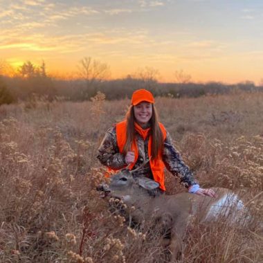 A woman poses at sunset in a field with a harvested deer.