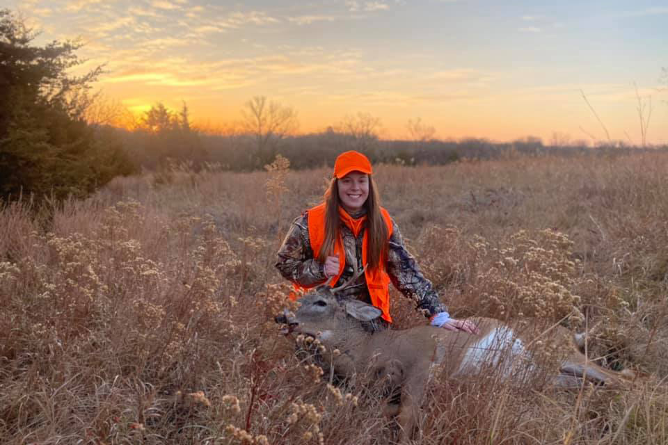 A woman poses at sunset in a field with a harvested deer.