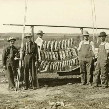 Black and white photo of men standing near stringers of fish.