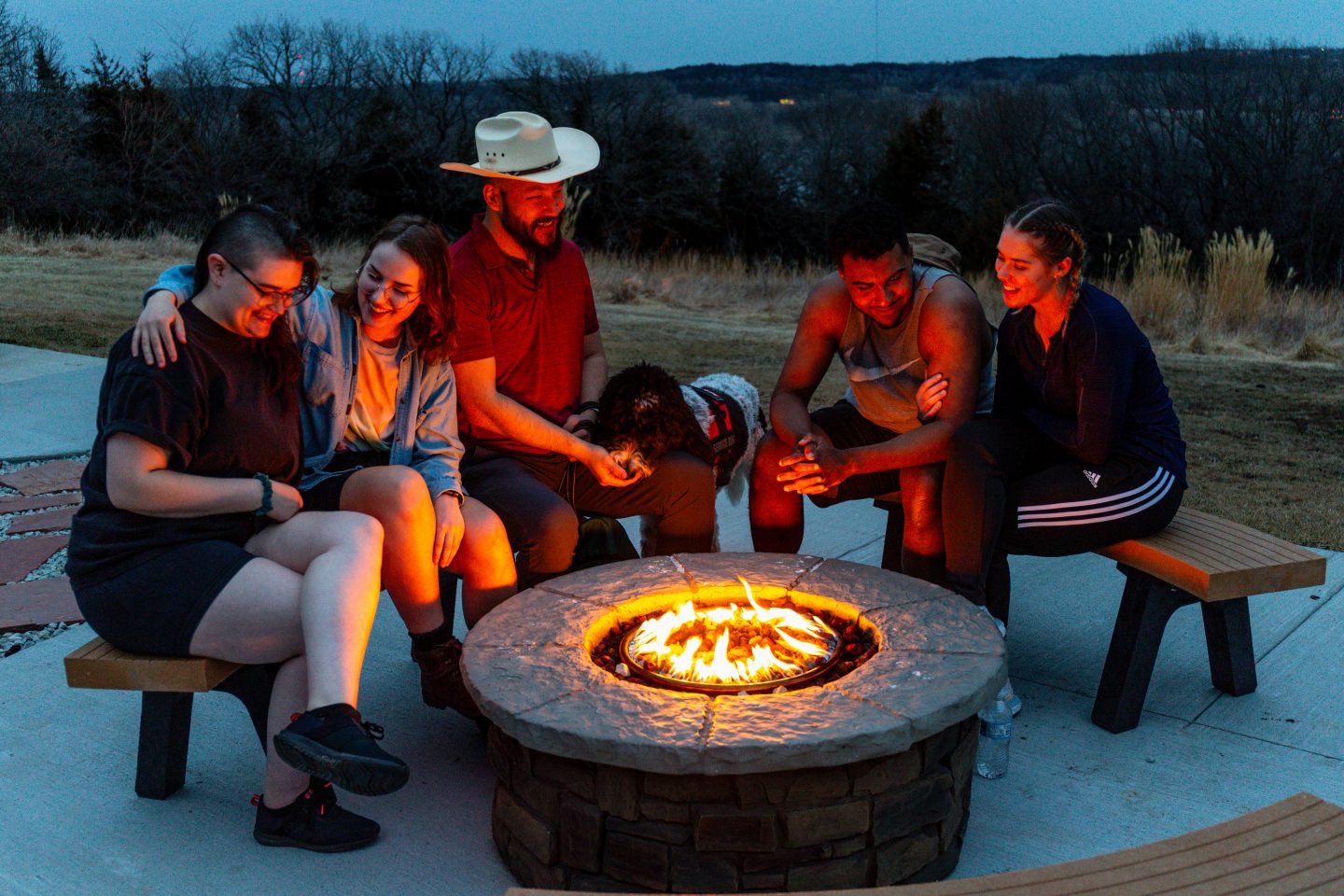 Ellie Piersol, Jamie Reigert, Francisco Grace, Alex Saw, and Cassidy Allen talk around a fire pit at Eugene T. Mahoney State Park in T-shirt and shorts weather.
