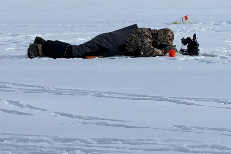 Angler laying on ice looking down ice hole.