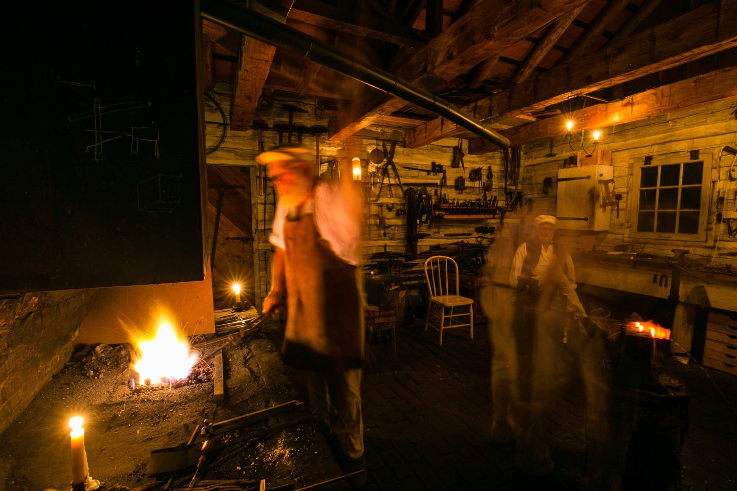 This photo, taken with a slow shutter speed during a Candlelight Tour, shows blacksmiths Doug Appel at the forge, Slader’s ghost-like form hammering at the anvil to the right and Lauren Joyce sitting at the work bench in the back.