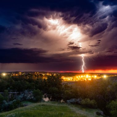 Lightning strikes in the distance over city lights from the vantage point of a hillside during summer.
