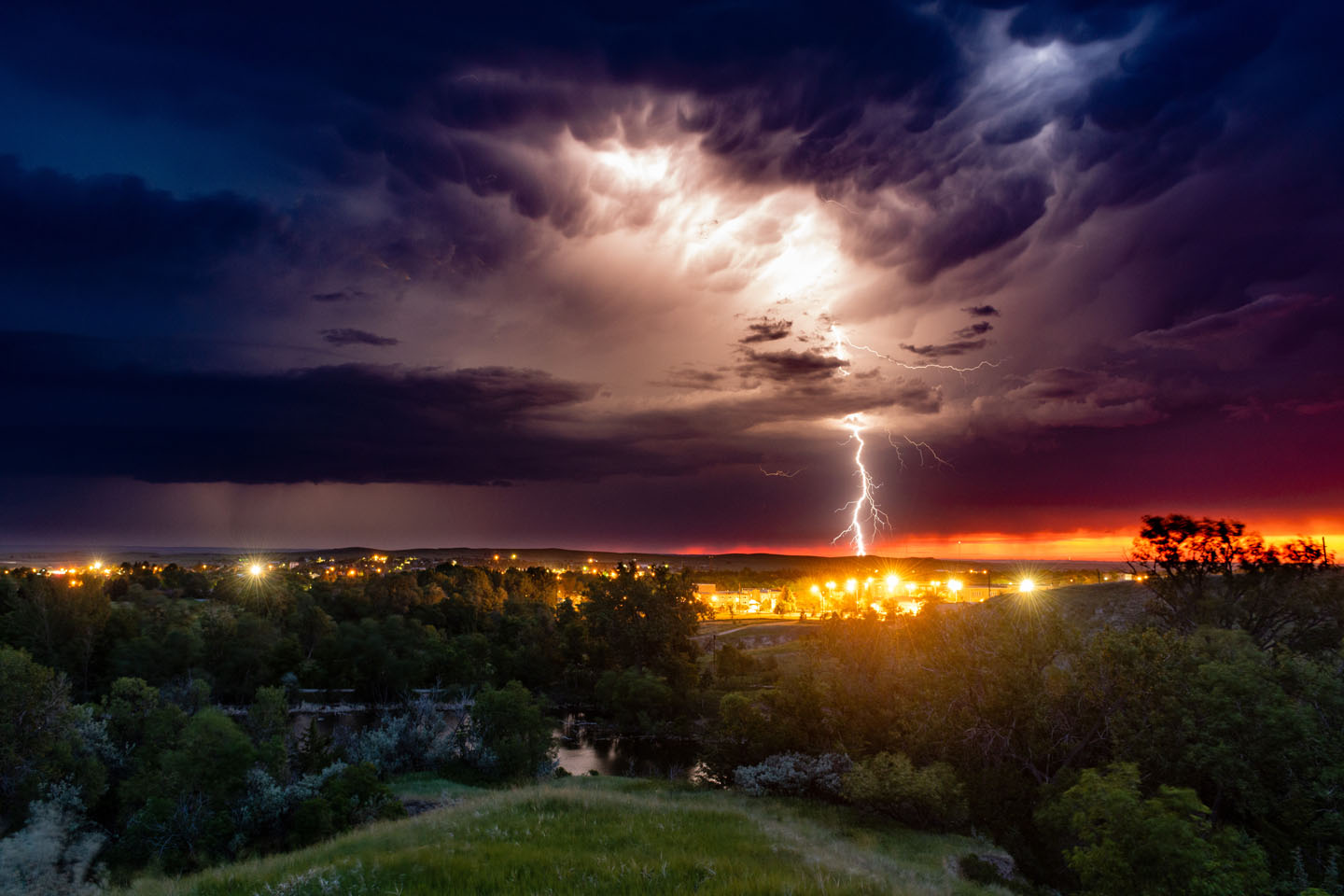 Lightning strikes in the distance over city lights from the vantage point of a hillside during summer.