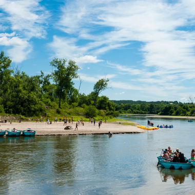 people in blue round vessels float past a sandy beach under bright blue skies