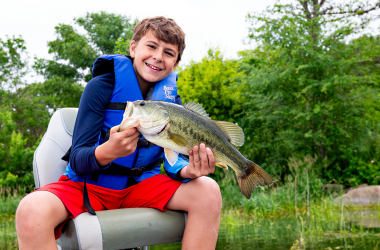 a boy in a blue life jacket holds a bass fish while sitting in a boat seat