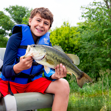 a boy in a blue life jacket holds a bass fish while sitting in a boat seat