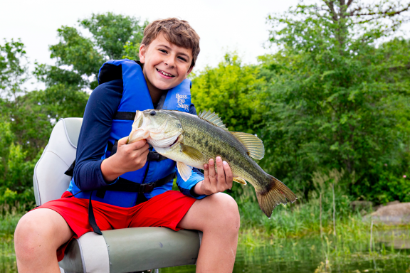 a boy in a blue life jacket holds a bass fish while sitting in a boat seat