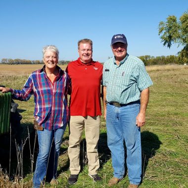 Three people stand by farm equipment in rural Nebraska.